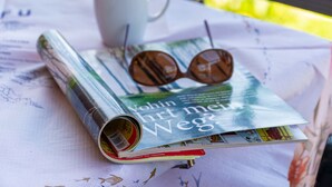 Un magazine, une paire de lunettes de soleil posées dessus, une tasse blanche et une table recouverte d’une nappe blanche.