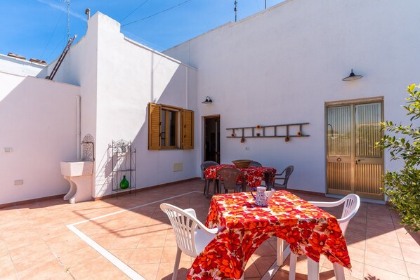 A patio with a table and chairs, a white wall, a wooden door, and a window with shutters.