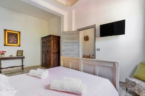 A bedroom with a bed, a wooden wardrobe, a bedside table with a vase and picture, a mirror, and a TV mounted on the wall.
