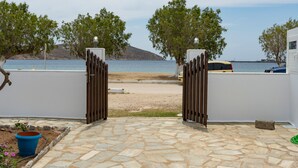 A stone-tiled patio leading to a beach with a white wall and a gate, trees, and a vehicle parked nearby.