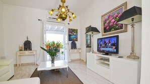 Olohuoneessa on valkoinen sohva, kukilla koristeltu sohvapöytä, valkoinen TV-taso, jossa on televisio, ja koristeellinen kattokruunu.