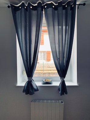 Janela com cortinas finas amarradas na parte inferior, um radiador abaixo e uma tigela no parapeito.