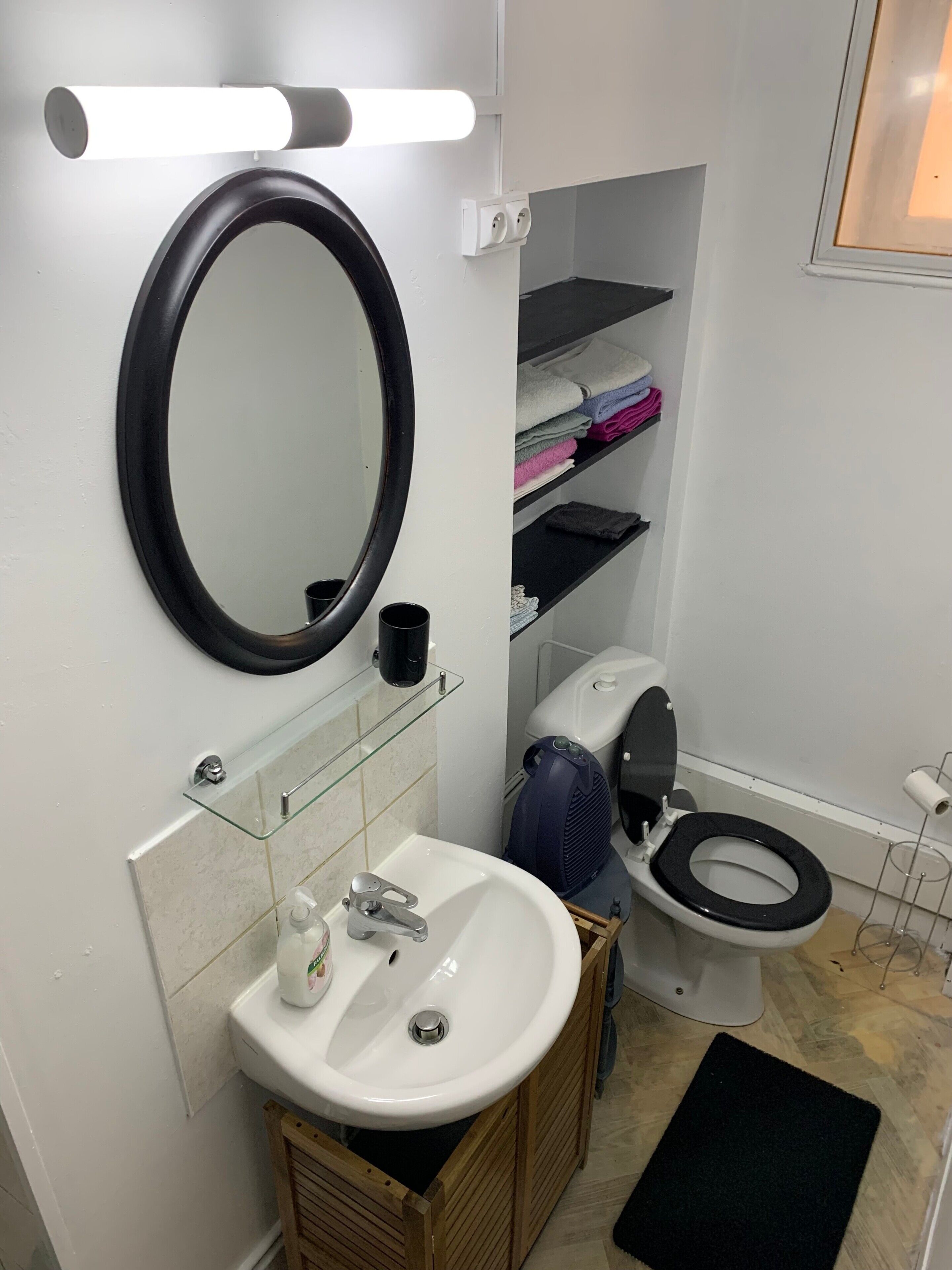 A bathroom with a white sink, a black toilet, a mirror, and a shelf with folded towels.