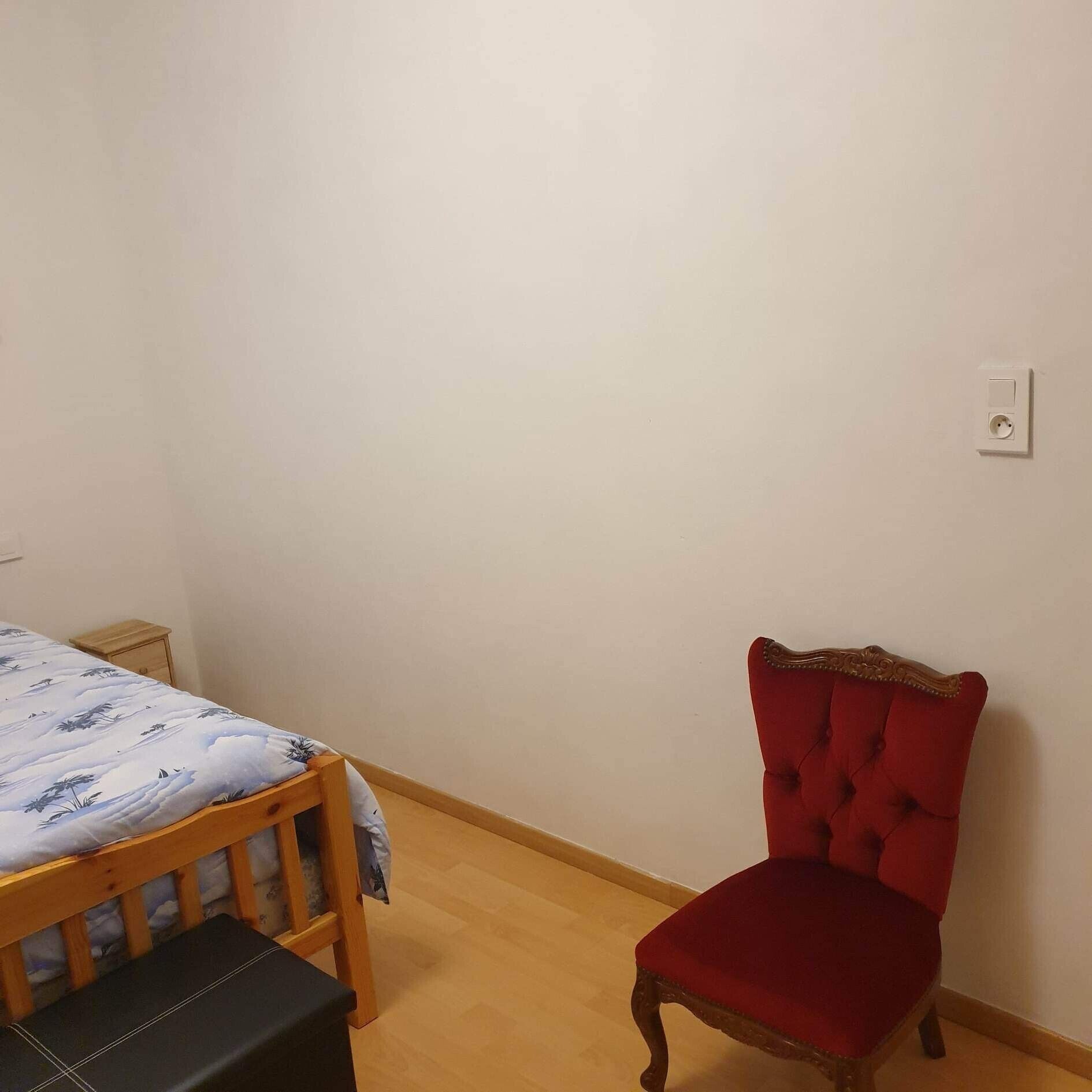 Een slaapkamer met een houten bed, een rode fauteuil, een nachtkastje en een stopcontact.