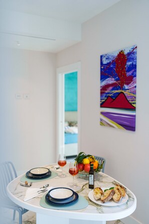 Une table à manger dressée avec des assiettes, des verres et une bouteille d’huile d’olive, ainsi qu’un panier de fruits et un tableau accroché au mur.