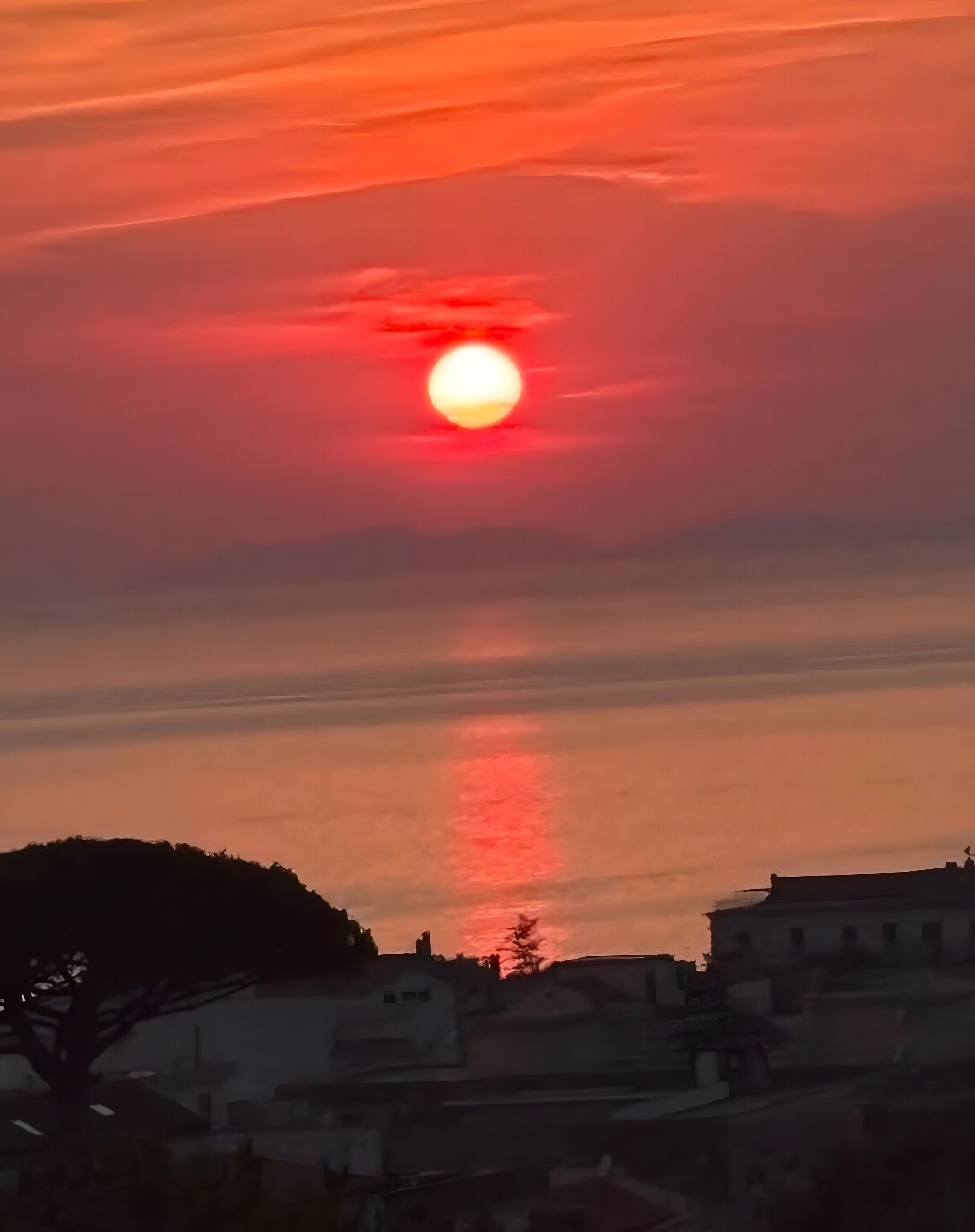 Un capvespre amb un sol visible, la silueta d'una collada i una filera d'edificis.