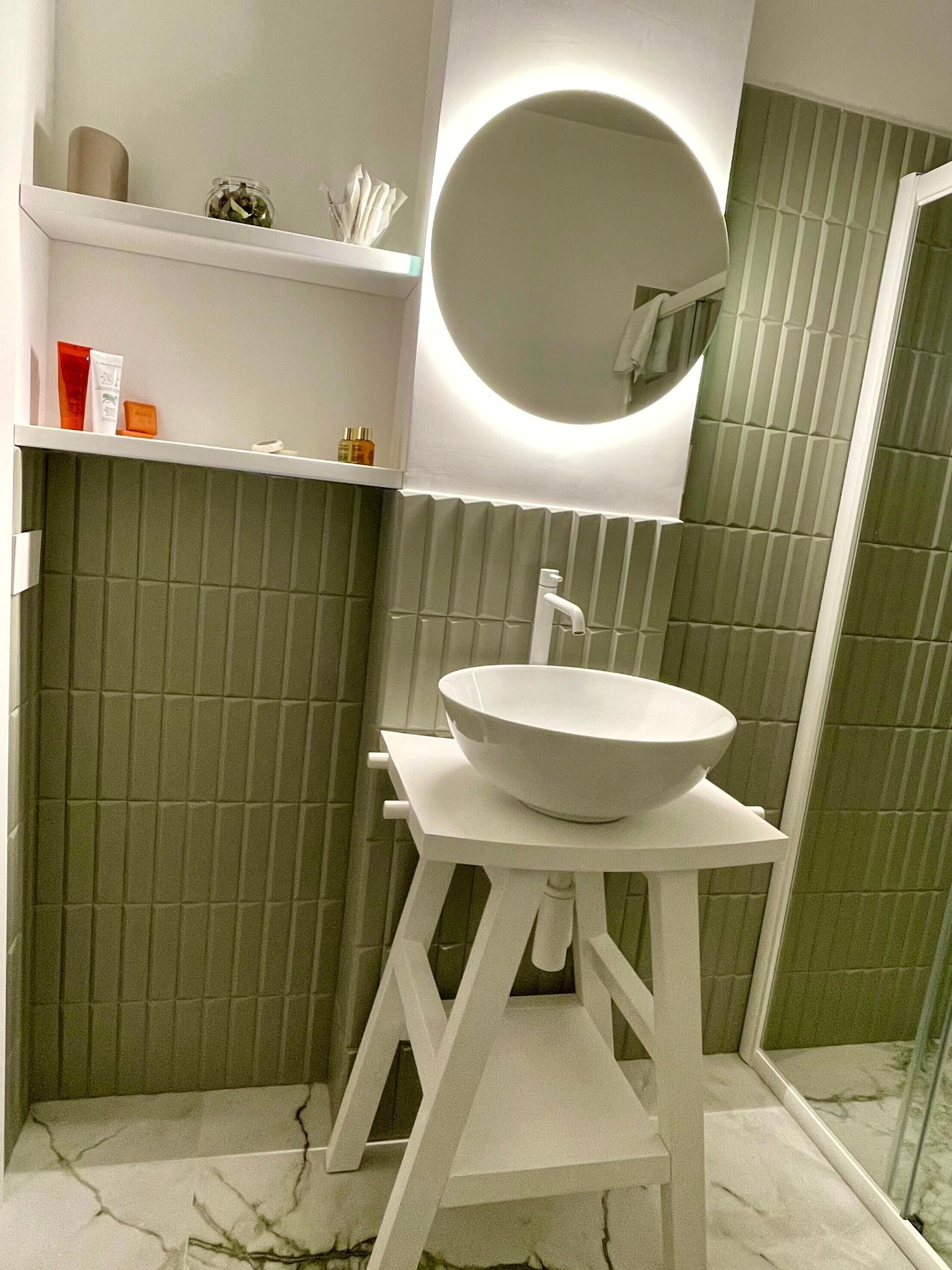 A bathroom with a white pedestal sink, a round mirror, and a shelf with toiletries.