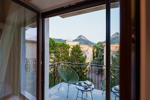A balcony with a view of buildings and mountains, a chair, and a table with a book and a cup.