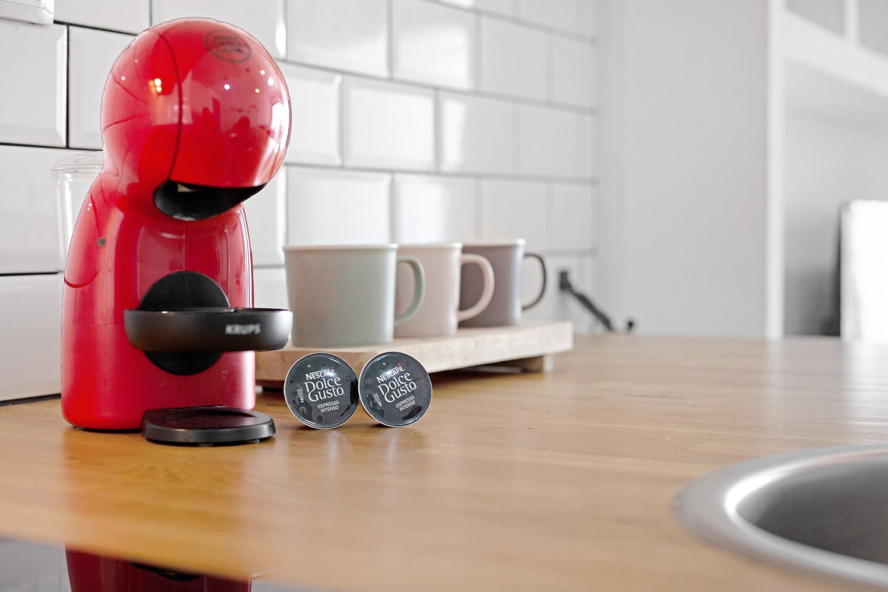 Een rode koffiemachine op een keukenblad met twee koffiekapsules met de tekst 'Nescafe Dolce Gusto Espresso' en een witte betegelde achterwand.