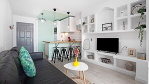 Un espacio habitable moderno con un sofá, una mesita redonda, un televisor y una zona de cocina con taburetes.