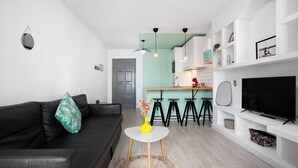 Una sala de estar moderna con un sofá negro, una mesita blanca con un florero amarillo, un televisor sobre un mueble blanco y una cocina al fondo.