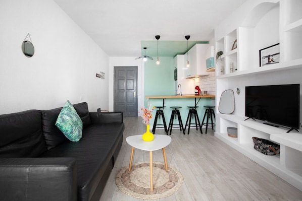 Ein modernes Wohnzimmer mit einem schwarzen Sofa, einem kleinen weißen Tisch mit einer gelben Vase, einem Fernseher auf einem weißen Schrank und einer Küchenzeile im Hintergrund.