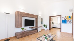 Una sala moderna con un televisor de pantalla plana, mueble de entretenimiento de madera, plantas en macetas y una mesa de centro de vidrio.
