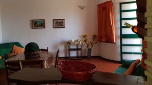 A cozy living room with a green sofa, a wooden table, and a red woven basket. There are two framed pictures on the wall, a small table with a vase of flowers, and a window with orange curtains.