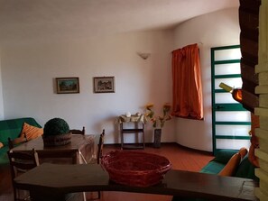 Una sala de estar acogedora con un sofá verde, una mesa de madera y una cesta trenzada roja. Hay dos cuadros enmarcados en la pared, una mesita con un jarrón de flores y una ventana con cortinas naranjas.
