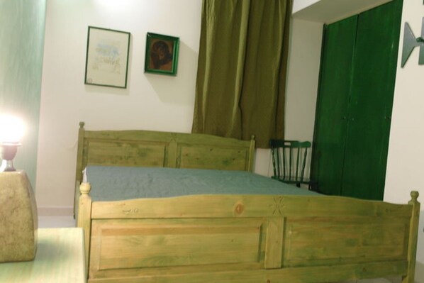 Una cama de madera con un colchón azul en una habitación con paredes blancas, una puerta verde y dos cuadros enmarcados.
