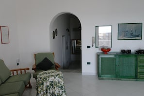 Ein Wohnzimmer mit Sofa, Sessel und einem kleinen Tisch. Ein grünes Schrank mit Spiegel, und ein Gemälde ist an der Wand zu sehen. Ein Bogen führt in einen weiteren Raum.