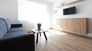 Un salon moderne comprenant un canapé, une petite table avec une plante, un téléviseur fixé à un mur en bois et une grande fenêtre ornée de rideaux légers.