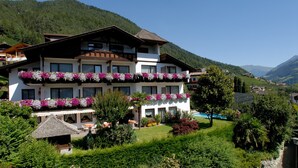 Um hotel com vários andares, varandas decoradas com flores, piscina e montanhas ao fundo.