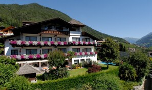 A multi-story hotel with balconies adorned with flowers, a pool, and a mountainous backdrop.