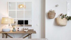 Ein Zimmer mit einem Holztisch, einer Lampe, einer Vase mit Blumen, einem 'Welcome'-Schild, einem Fenster mit einer Pendelleuchte und zwei wandmontierten Flechtkörben.