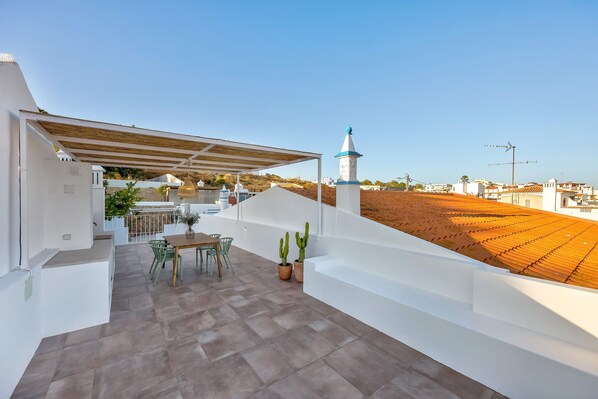 A rooftop terrace with a pergola, a table with chairs, potted plants, and a view of the surrounding landscape.