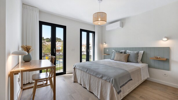 Ein Schlafzimmer mit einem Bett, einem Schreibtisch und einem Stuhl. Ein Fenster bietet Blick auf Gebäude, und eine Tür führt in einen Außenbereich.