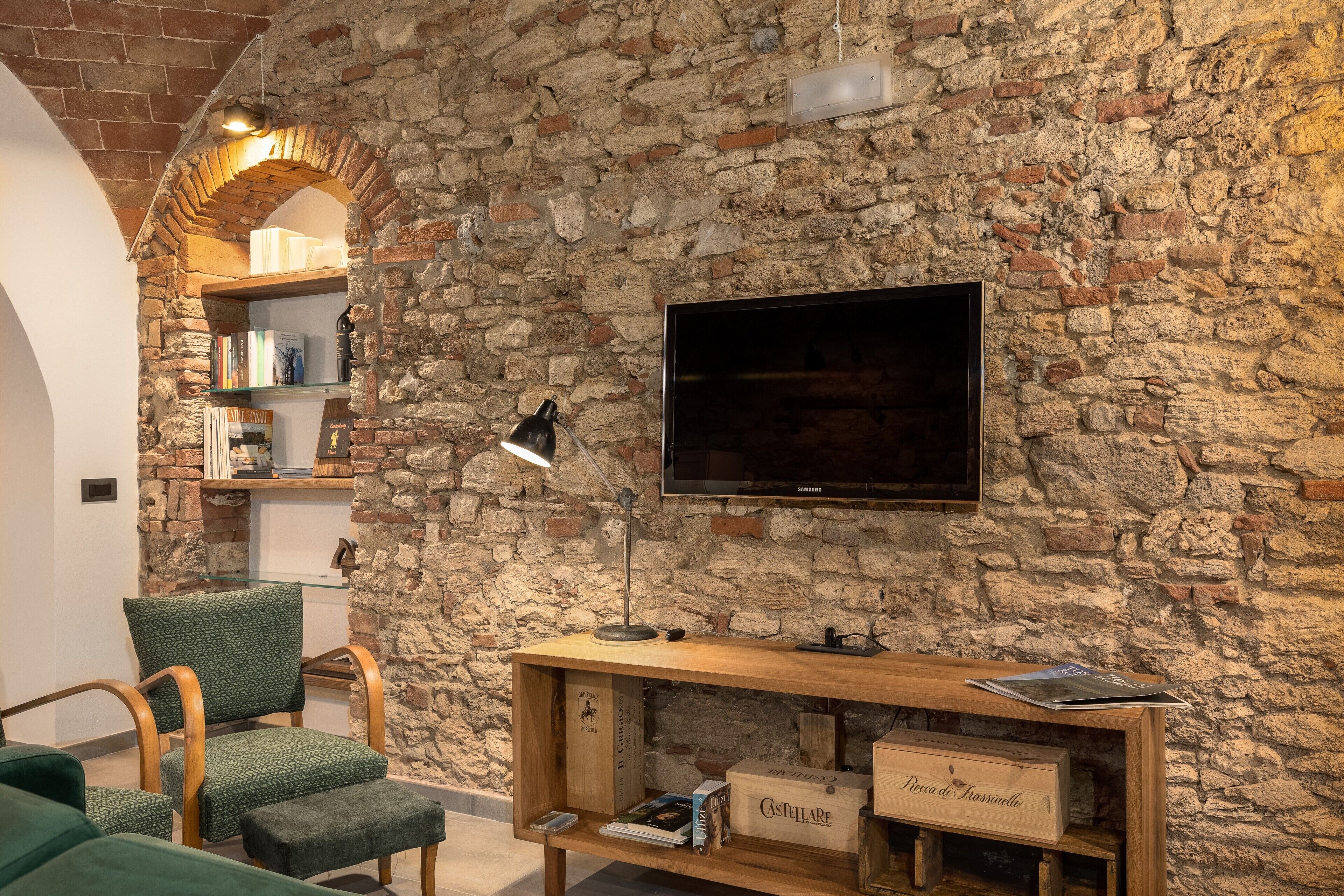 A room with a stone wall, a flat-screen TV, a wooden console table, a green upholstered chair, and a bookshelf.