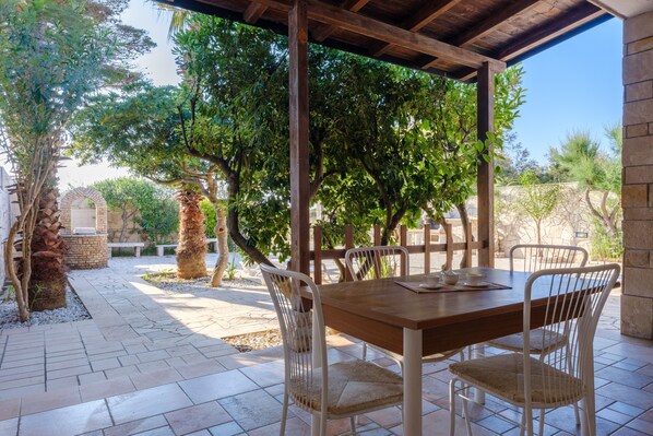 An outdoor patio with a wooden table and white chairs, surrounded by trees and a stone wall.