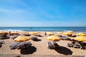 Une plage avec des transats et des parasols jaunes, une terrasse en bois et l’océan en arrière-plan.