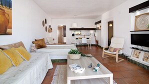 Un salon avec un canapé, une table basse et des fauteuils, un coin repas avec des chaises et une table, et une cuisine à l’arrière-plan.