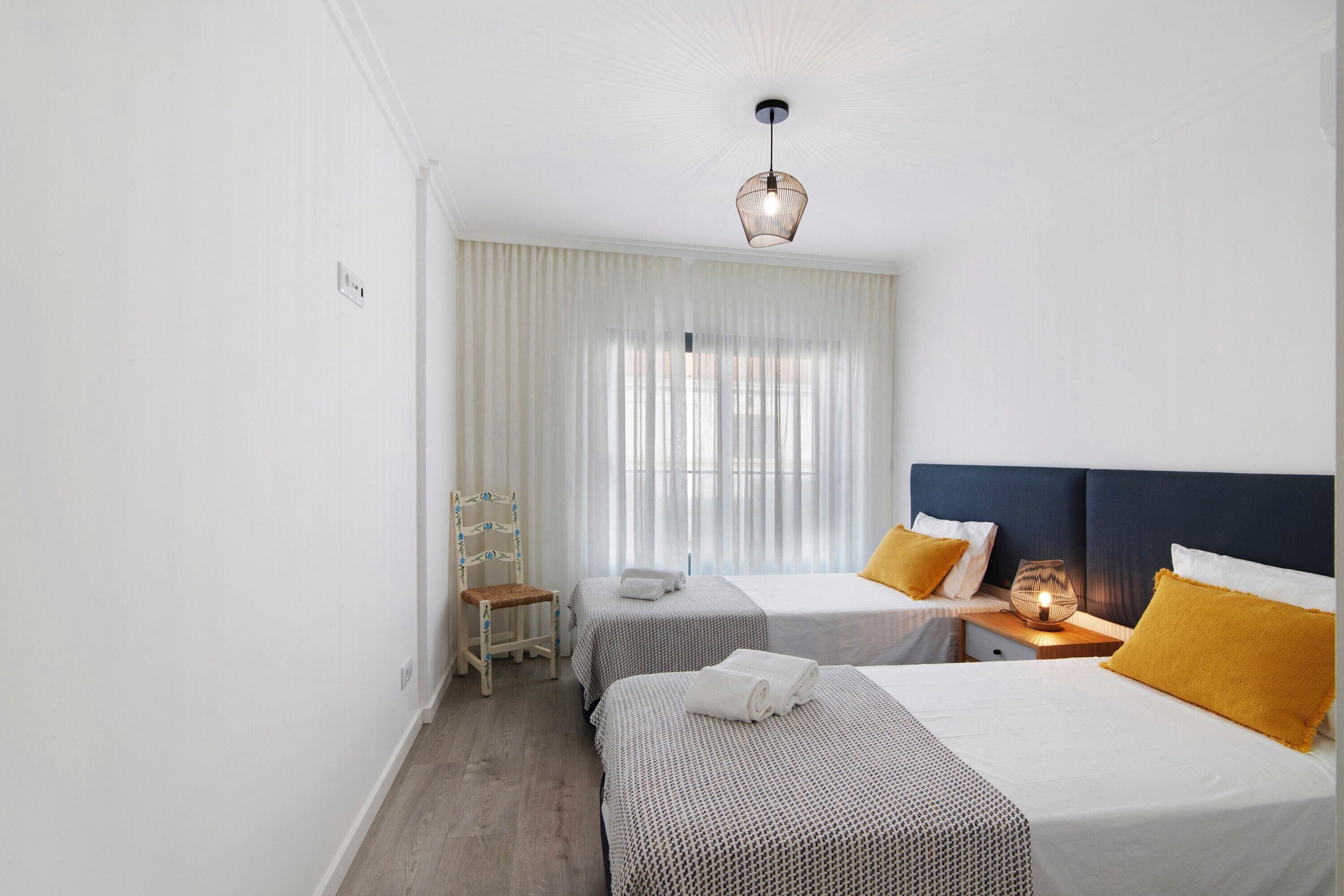 Habitación de hotel con dos camas, una silla, una mesita de noche con lámpara y un ventanal con cortinas.