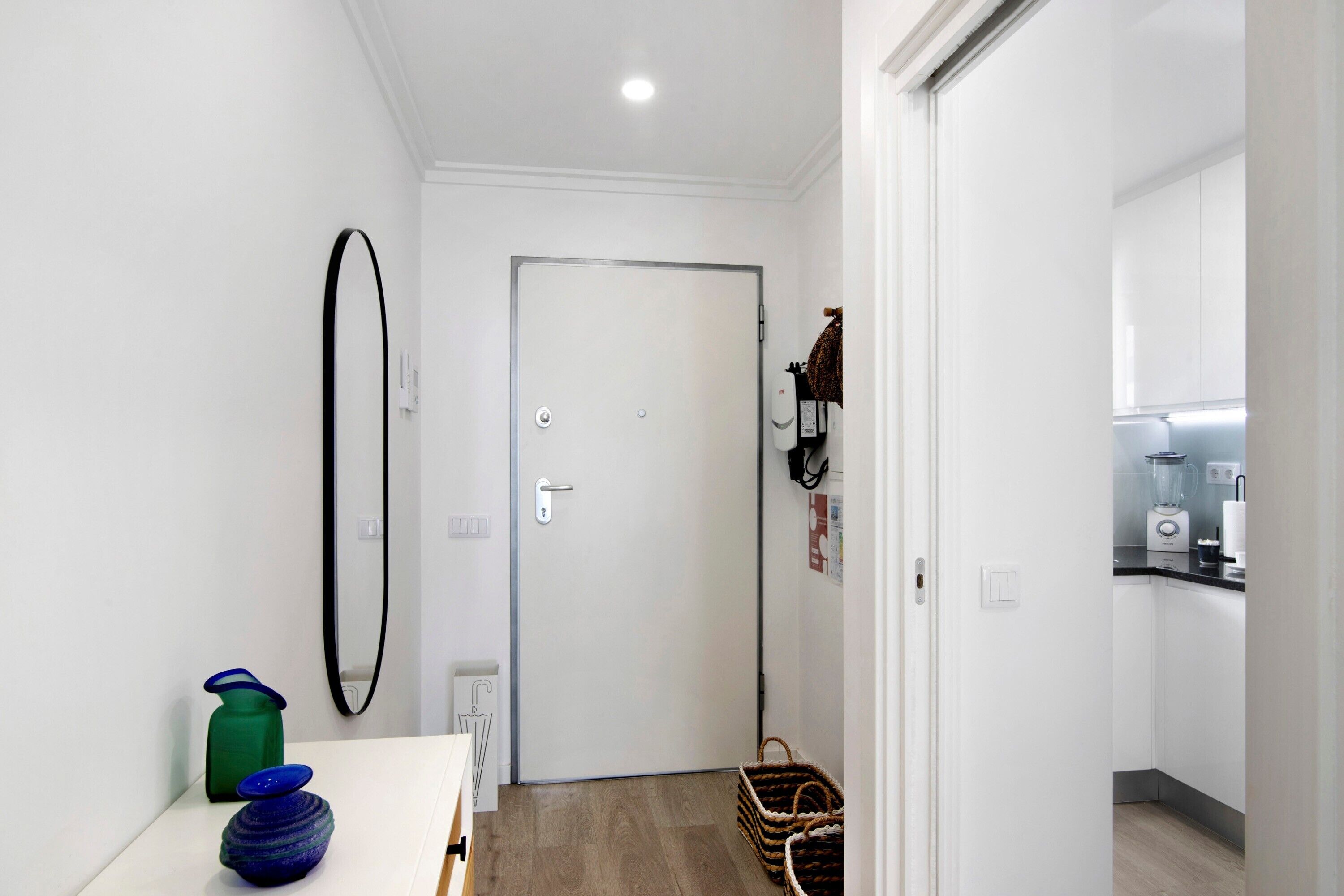 Un pasillo que lleva a una puerta blanca, una zona de cocina con una licuadora y una cesta de mimbre en el suelo.