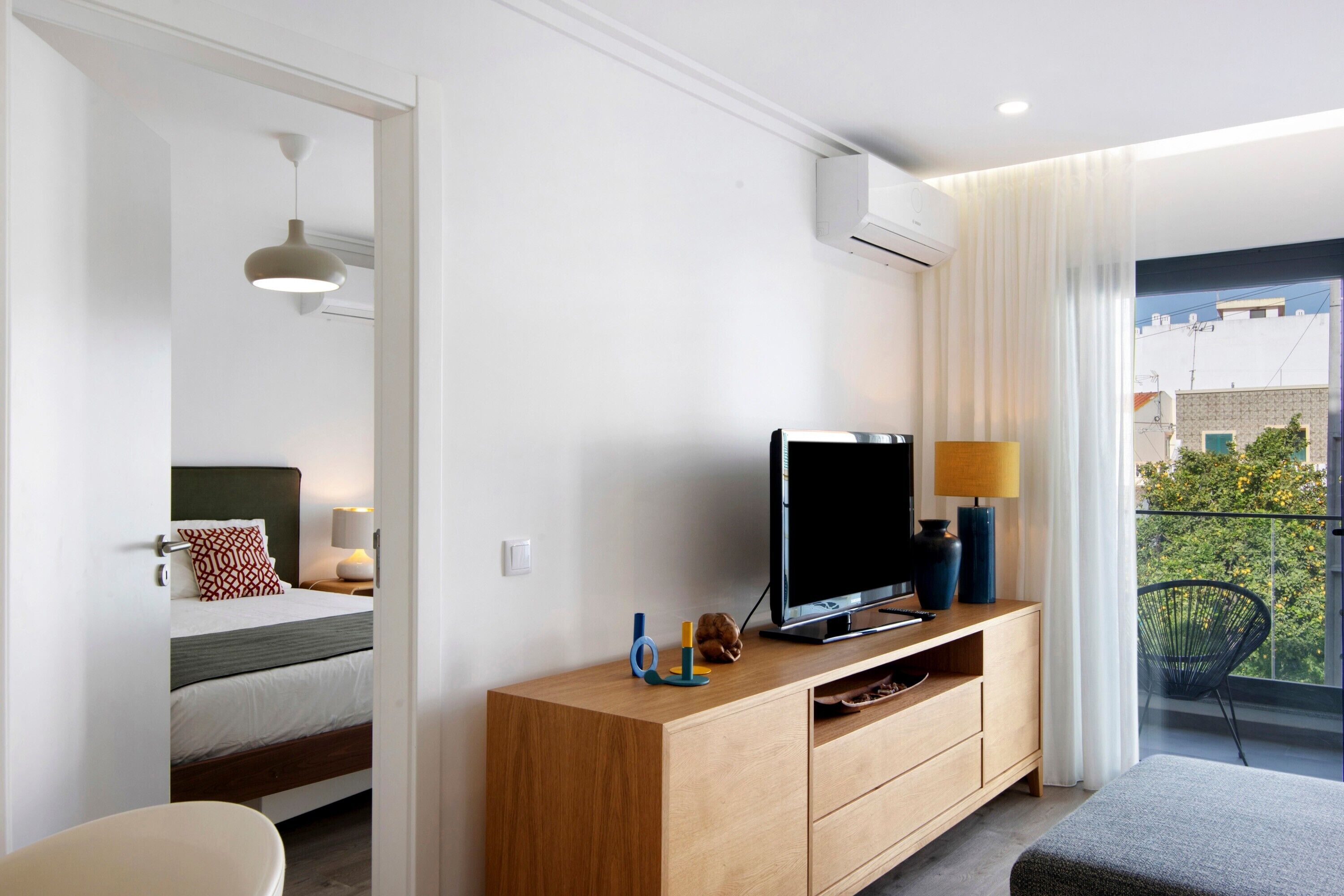 Una habitación de hotel moderna con cama, televisor sobre una consola de madera, balcón con vistas y unidad de aire acondicionado en la pared.
