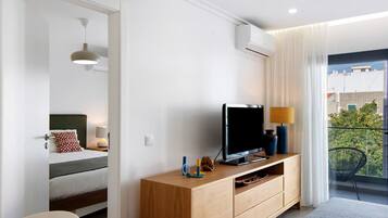 Una habitación de hotel moderna con cama, televisor sobre una consola de madera, balcón con vistas y unidad de aire acondicionado en la pared.