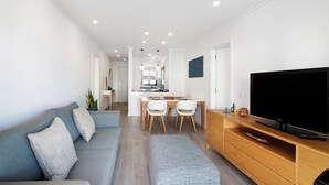En moderne stue med en grå sofa, et fjernsyn på et træmøbel, et spiseområde med bord og stole, og et køkken i baggrunden.