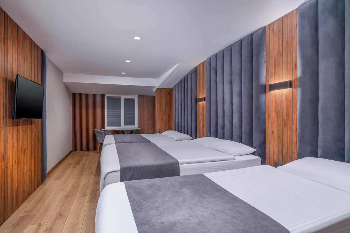 Une chambre d’hôtel avec trois lits, un téléviseur à écran plat, des murs lambrissés et une fenêtre avec des stores.
