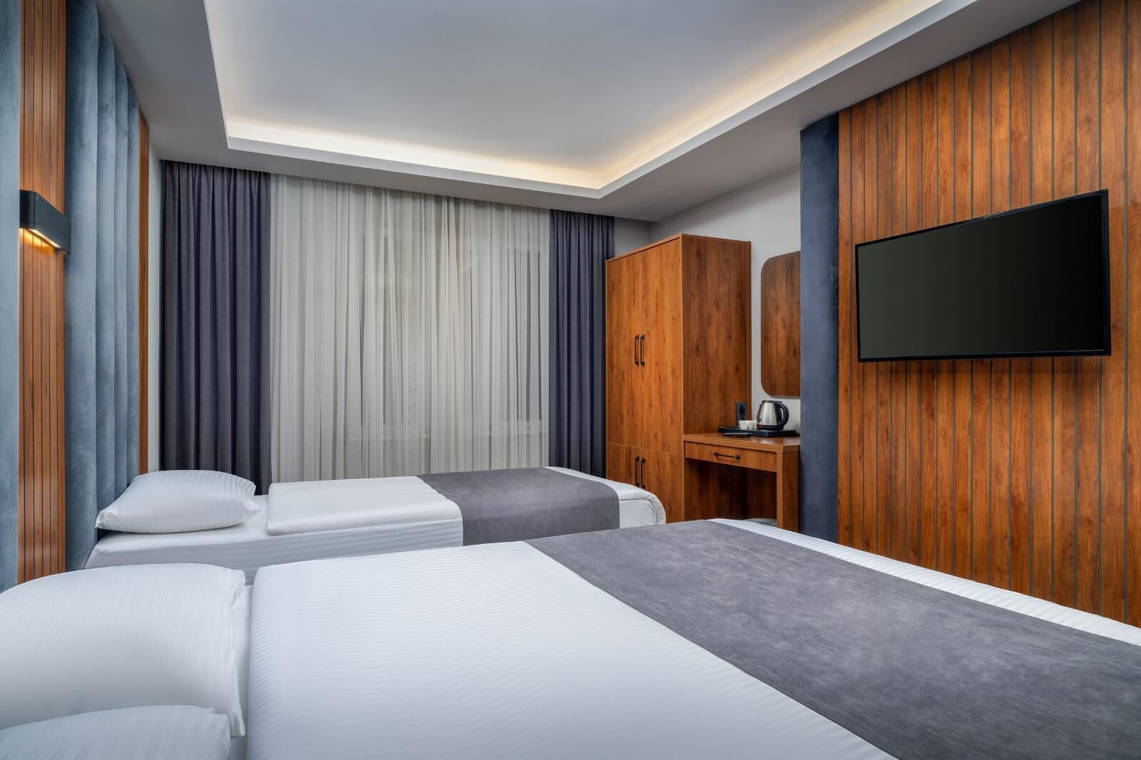 Hotellihuone, jossa on kaksi sänkyä, puinen vaatekaappi, peilillä varustettu työpöytä ja seinälle asennettu litteänäyttöinen televisio.