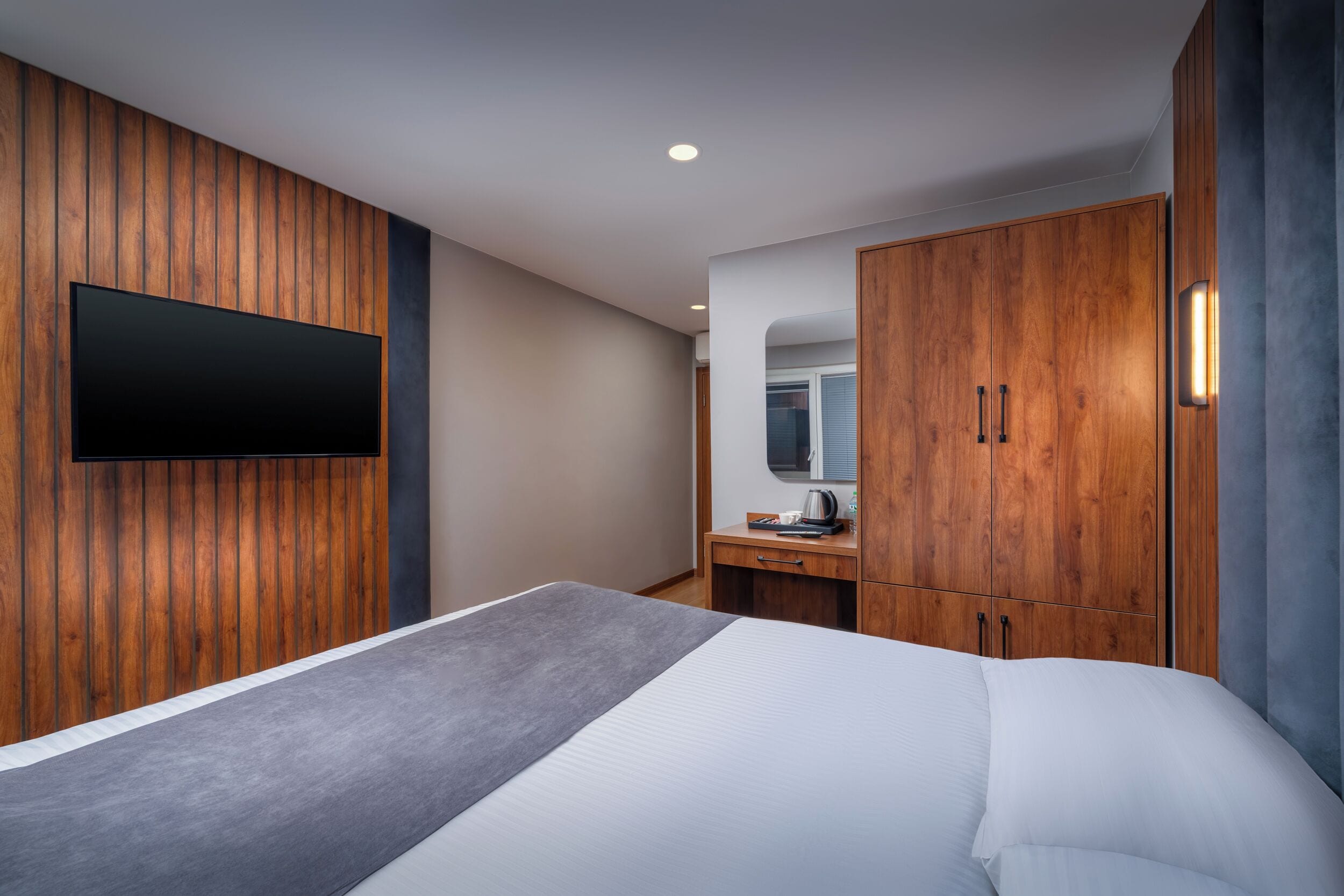 A modern hotel room with a large bed, wooden panel walls, a flat-screen TV, a wooden wardrobe, and a small desk with a mirror.