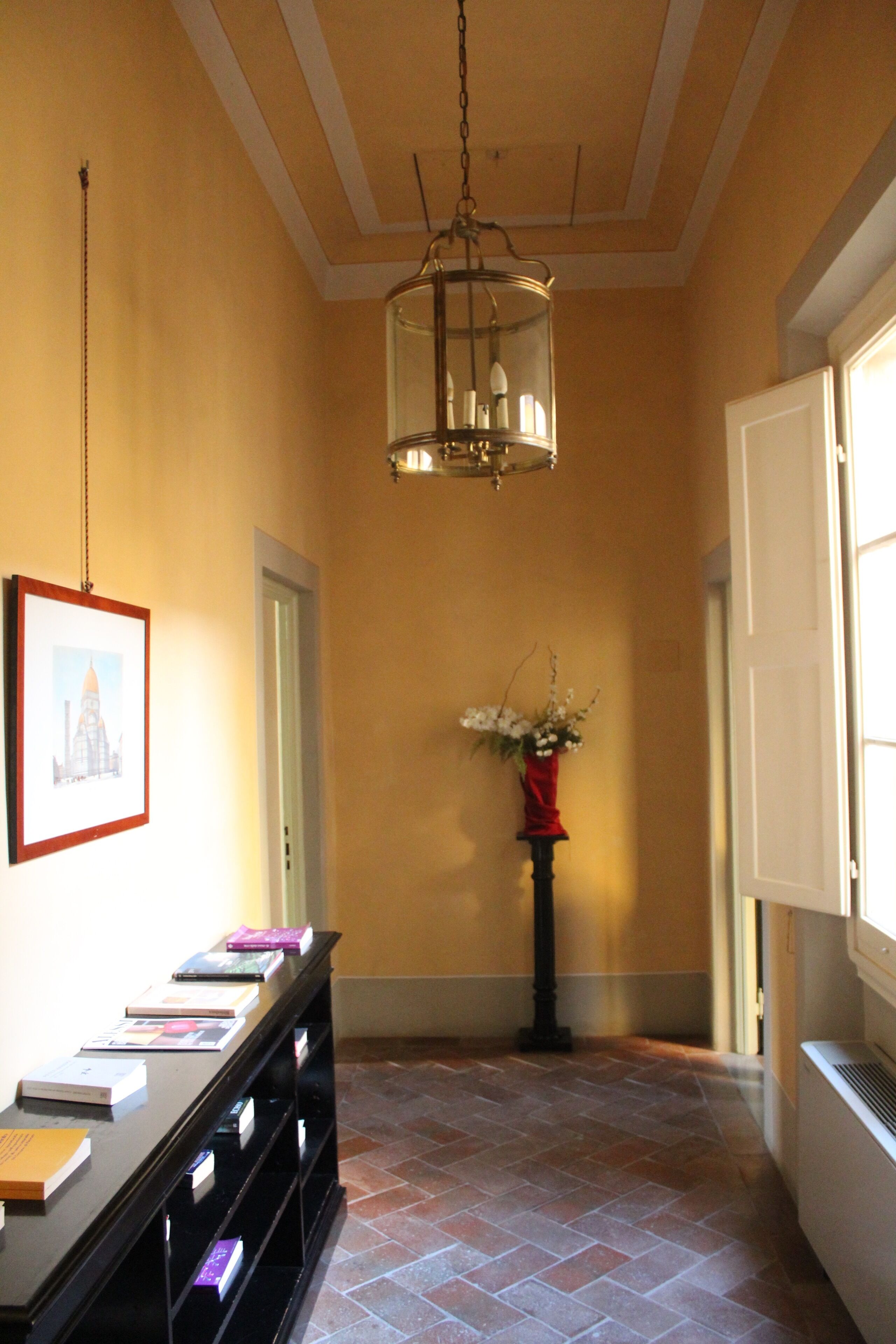 Um corredor com uma mesa consola preta exibindo livros, um quadro na parede, um vaso com flores e um candeeiro suspenso.