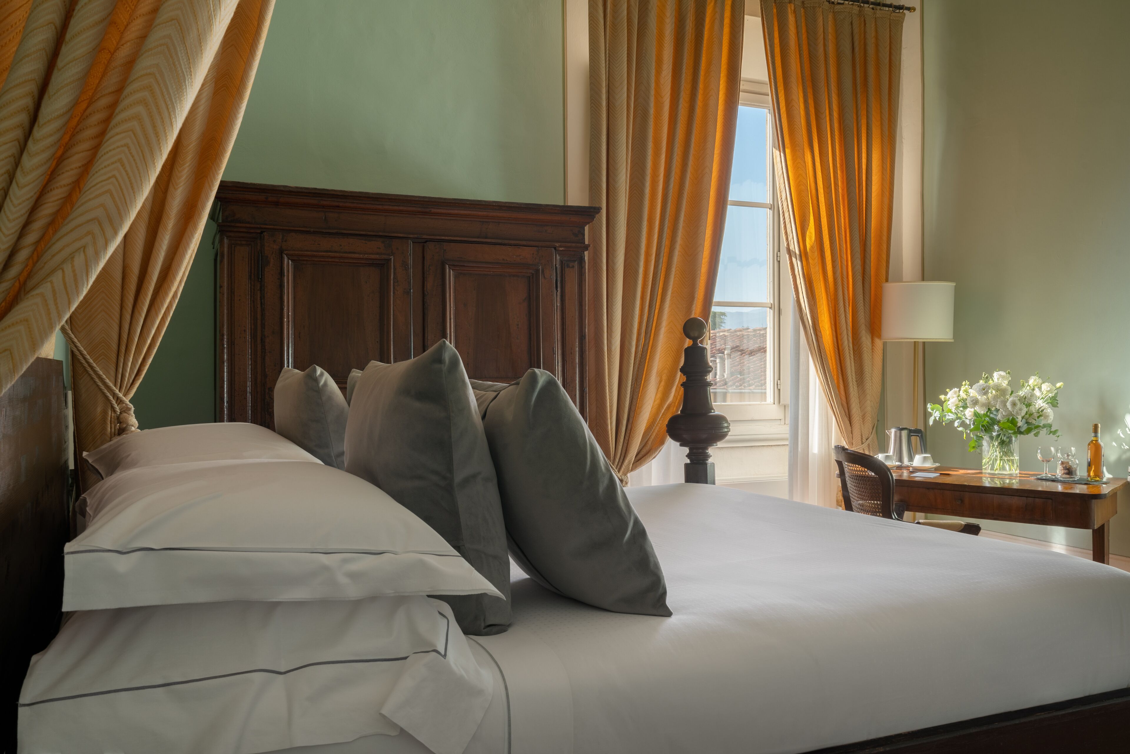 Una cama bien hecha con múltiples almohadas, un cabecero de madera y una mesita con un florero y una botella.