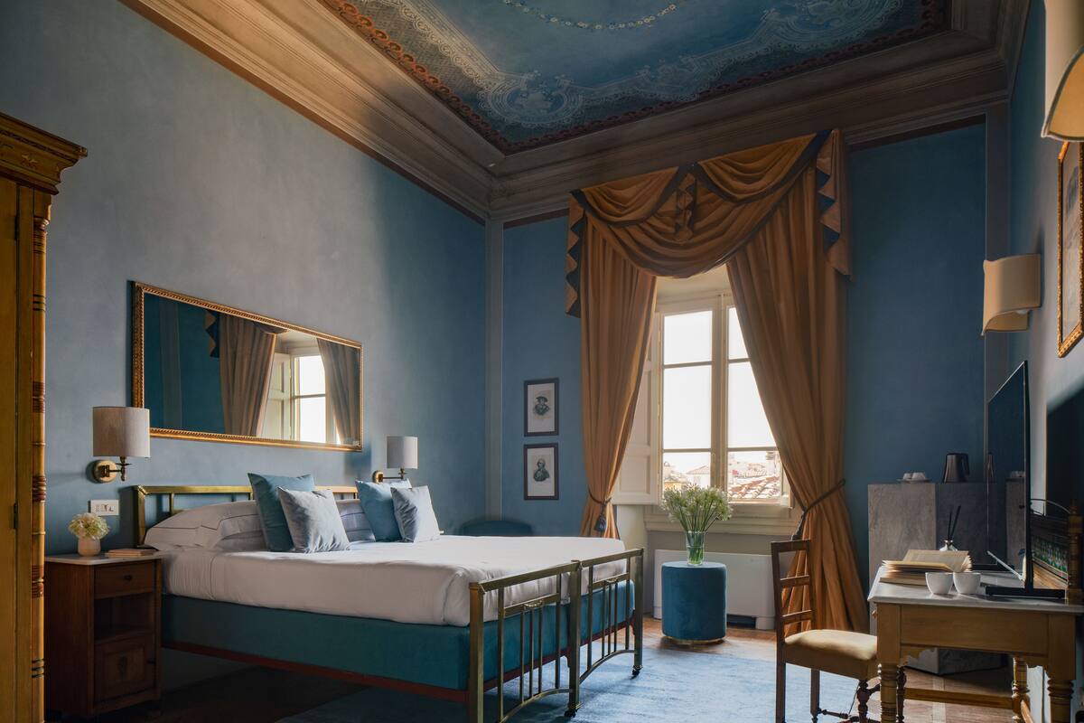 Une chambre à coucher comprenant un lit, une table de chevet, une lampe, un miroir, une fenêtre avec des rideaux et un bureau avec une chaise.