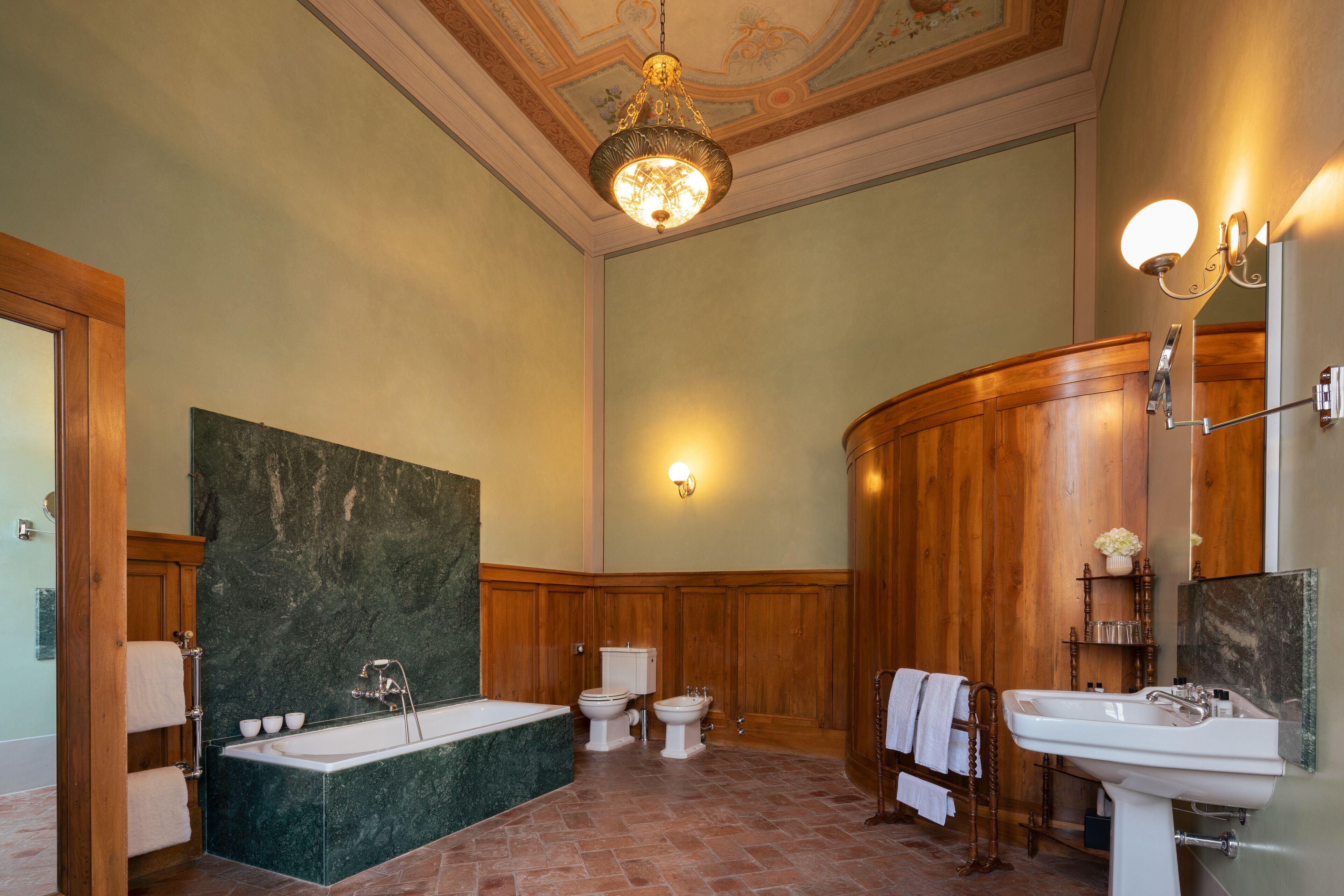 A bathroom with a bathtub, two toilets, a sink, wooden paneling, a chandelier, and a marble wall.