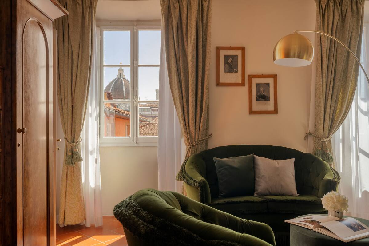 Une pièce avec un canapé vert, une porte en bois, une fenêtre donnant sur des immeubles et un lampadaire.