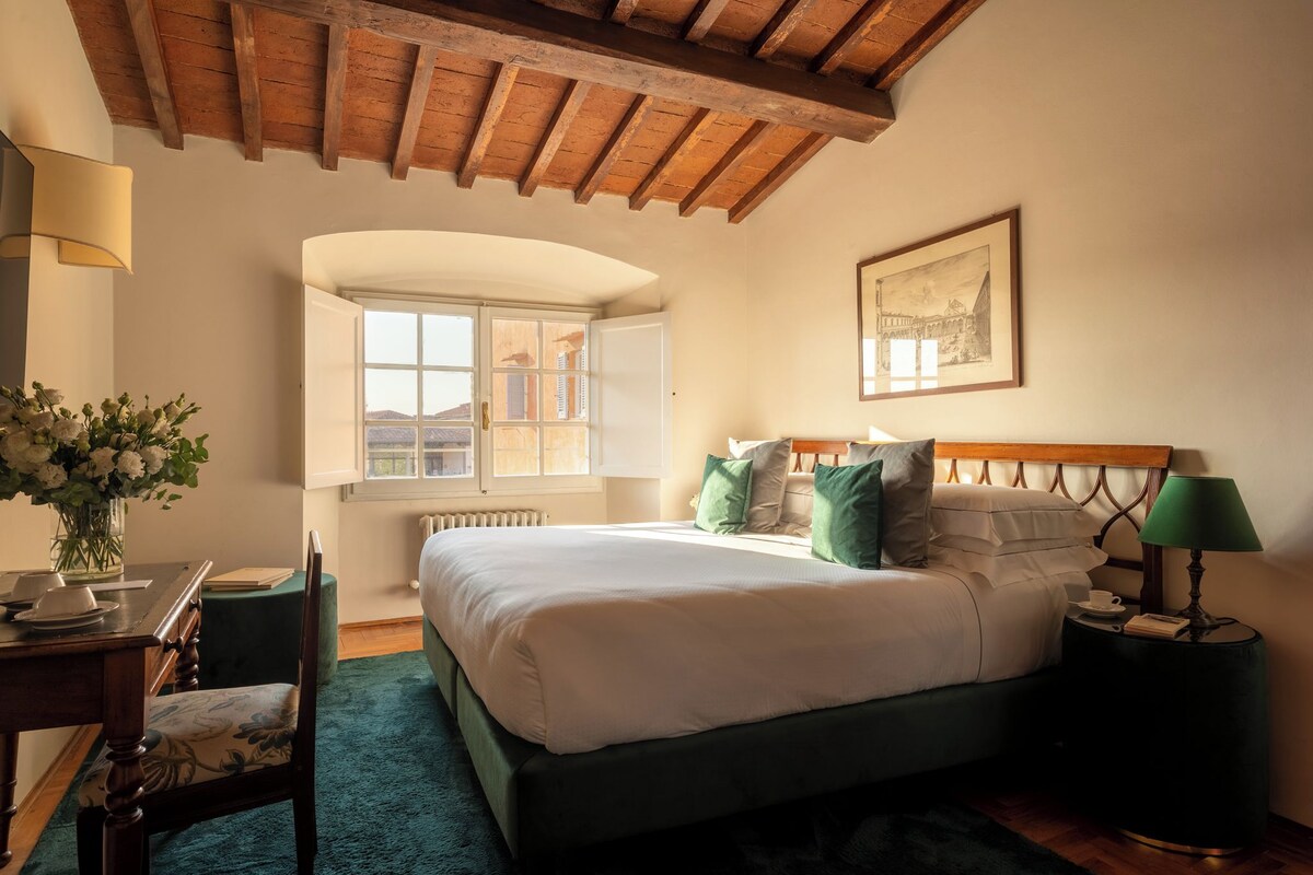 A bedroom with a large bed, a desk with a chair, a window, and a framed picture on the wall.