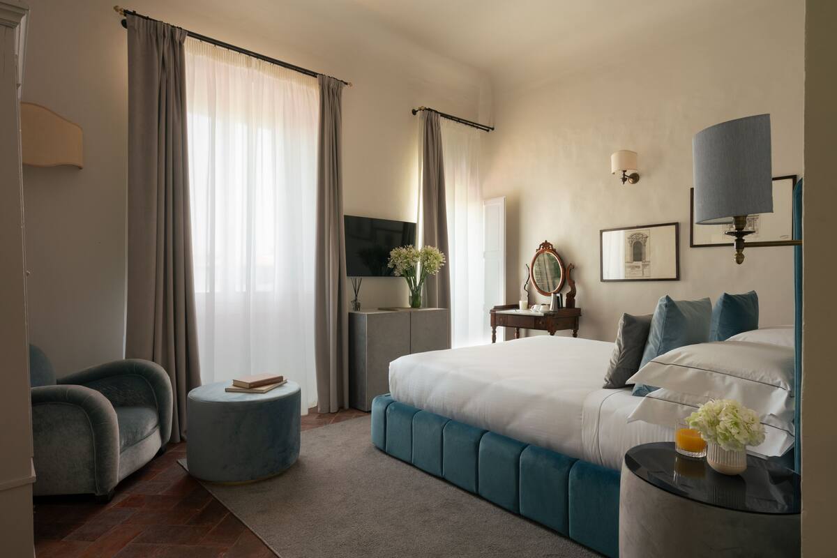 Une chambre d’hôtel moderne dotée d’un grand lit, d’un coin salon avec un fauteuil et un pouf, d’une coiffeuse avec un miroir et d’un téléviseur à écran plat.