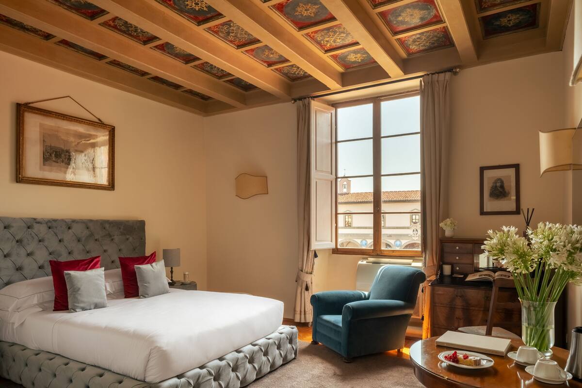 Une chambre d’hôtel avec un grand lit, un fauteuil bleu, un bureau avec une lampe et une vue sur un immeuble par la fenêtre.