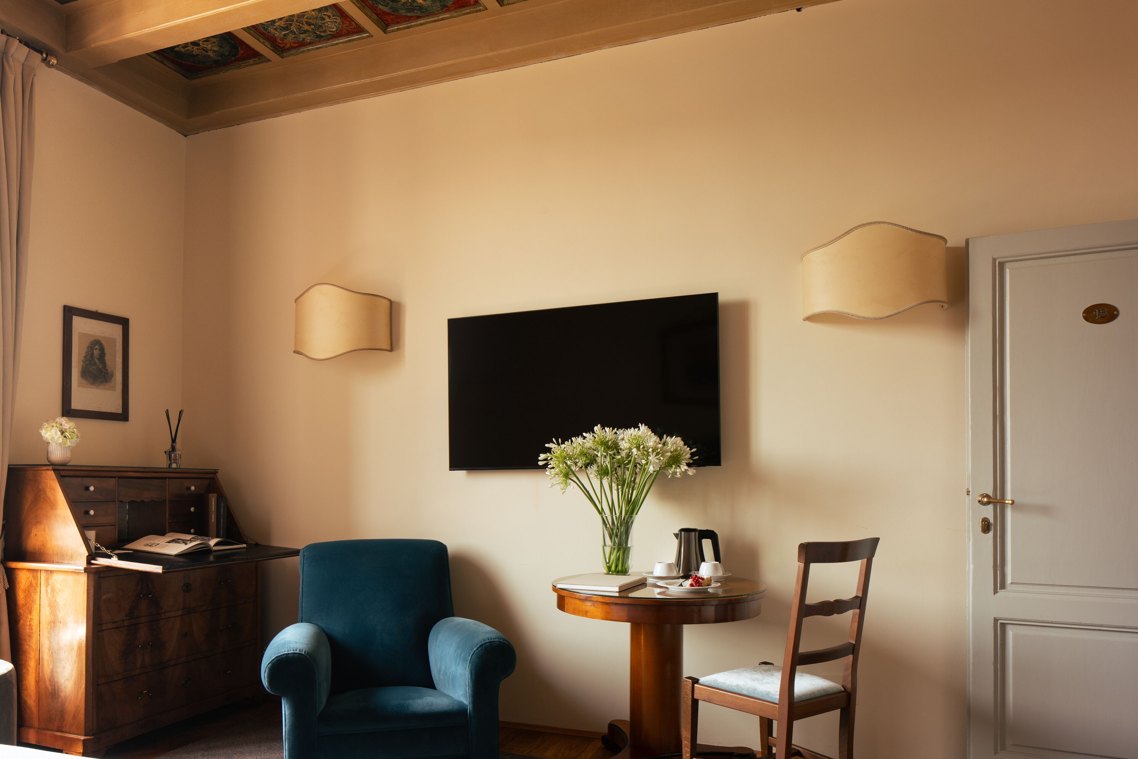 A room with a blue armchair, a wooden side table with a vase of flowers, a flat-screen TV, and a door with a brass handle.