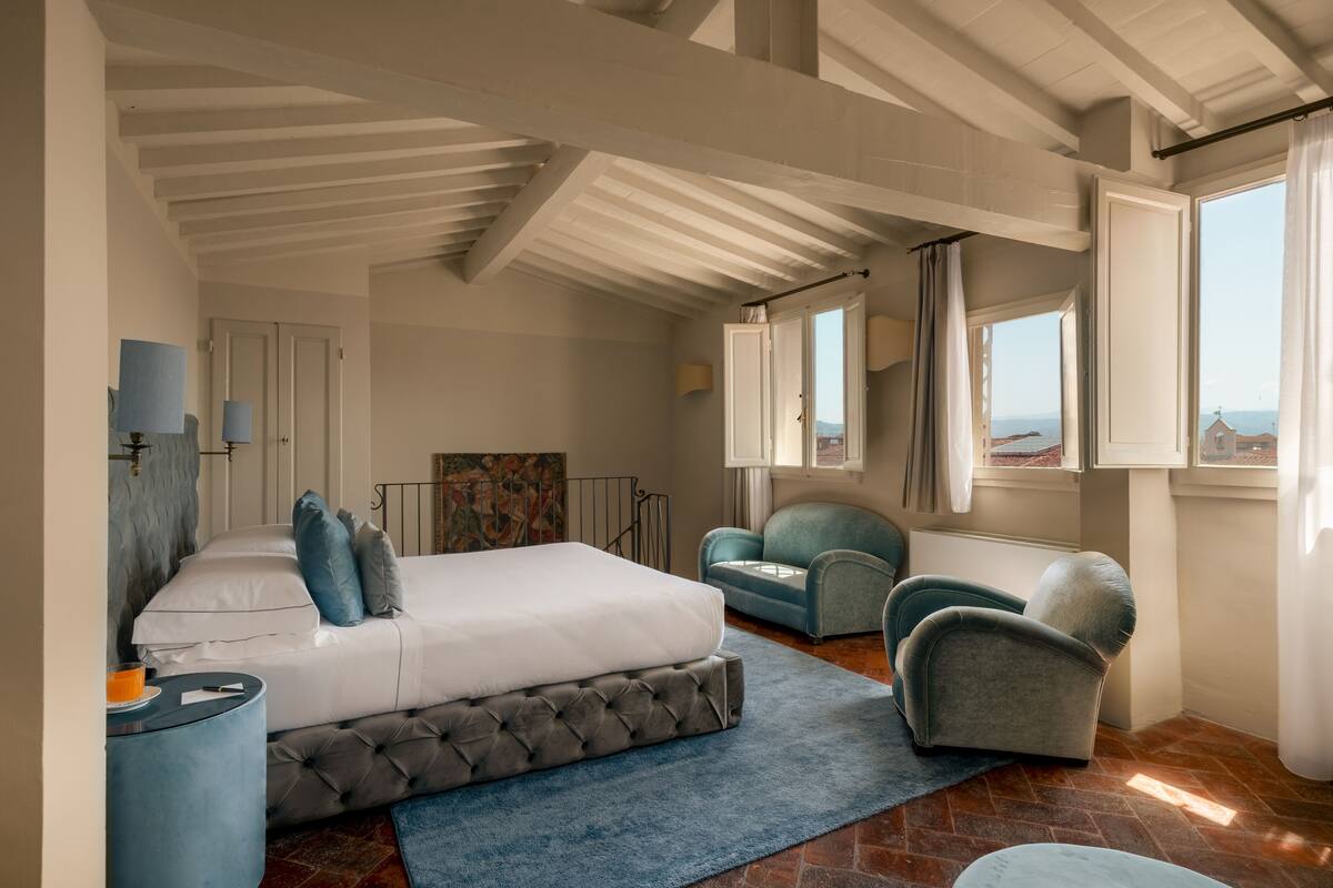Une chambre avec un grand lit, deux fauteuils, un tapis bleu et une vue sur la mer par les fenêtres.