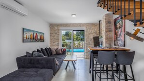 A modern living room with a gray sofa, a coffee table, and a dining area with bar stools. There is a stone wall and a large window overlooking a pool. - Three bedroom villa with pool (Malinska)